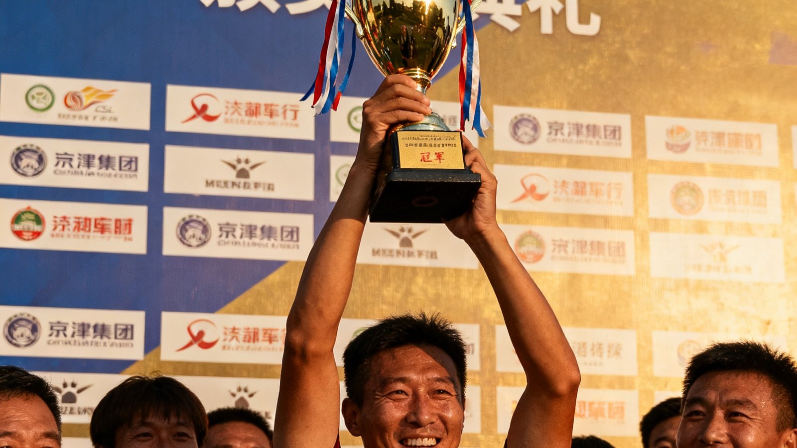2024年京津冀企业足球联赛颁奖典礼，获奖队伍高举奖杯，背景是赛事主视觉墙和赞助商logo。